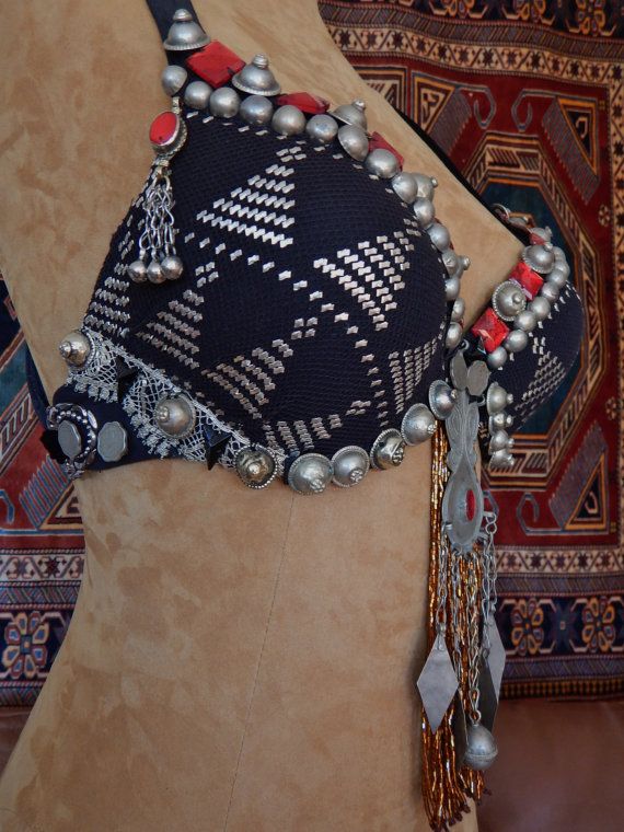 this is a closeup photo of a belly dance costume, showing the top, which is black and intricately embroidered. the top is covered with silver jewelry, red tassels, and is worn over a beige torso. the background is a closeup of a rug.