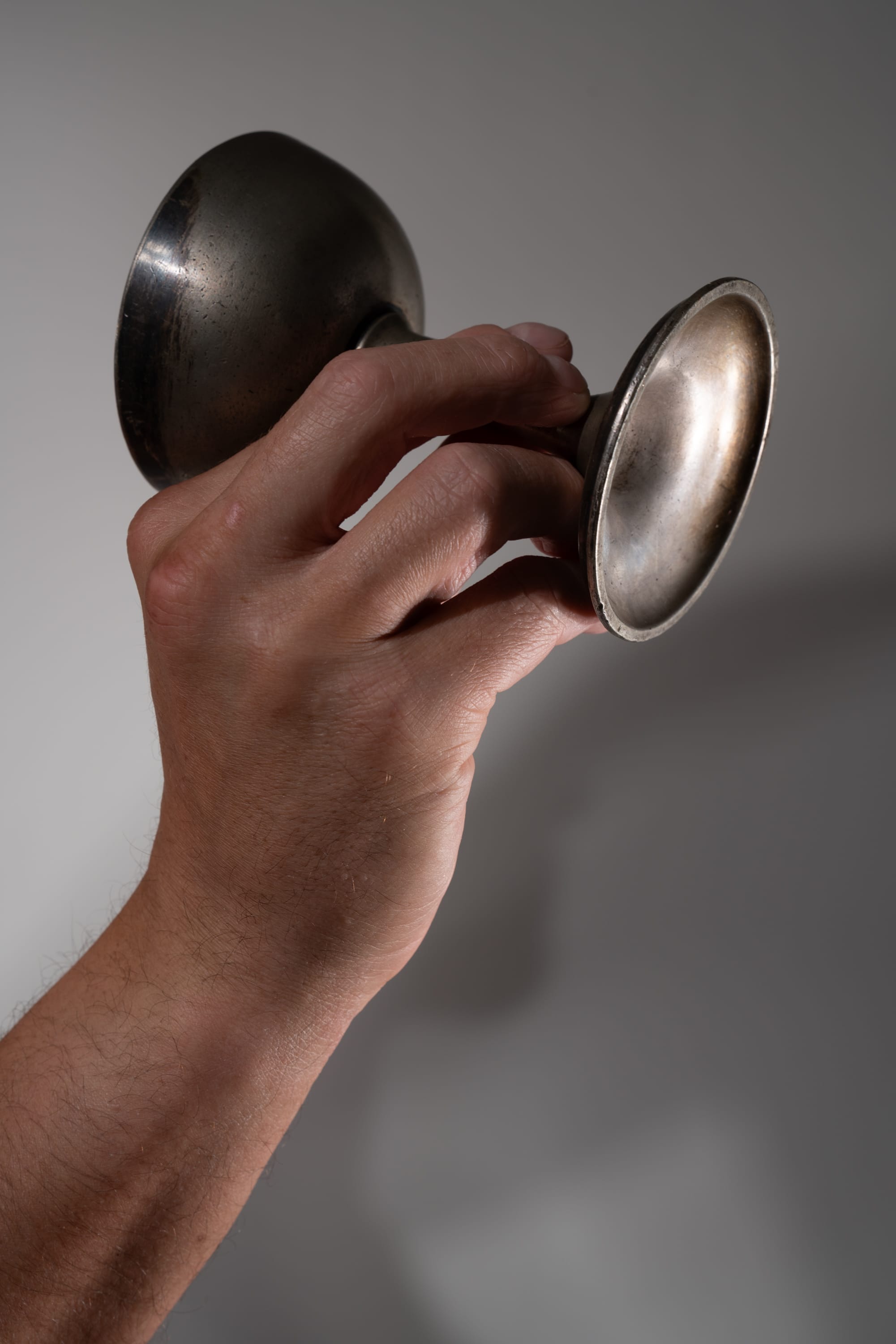 a closeup of a man's hand holding up a tarnished metal trophy against a white background. the trophy is made of a dull, silvery metal and has a rounded cup and a wide base. the hand is holding the trophy by the stem and base, and the fingers are curled around the base. the hand is illuminated by a strong light source from the left side, creating deep shadows on the right side of the trophy and hand. the image has a sense of pride and accomplishment, as the hand is displaying the trophy prominently.