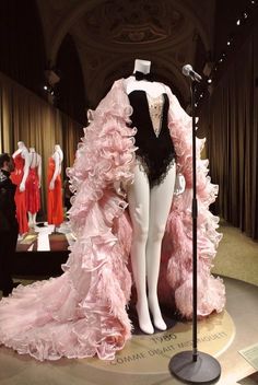 a captivating display of a vintage 1980s haute couture dress in a museum setting. the dress, a striking combination of black and pale pink, features a fitted black bodysuit with a sweetheart neckline and a dramatic floorlength cape crafted from layers of delicate pink ruffles.  the mannequin stands on a platform, illuminated by spotlights, creating a sense of drama and highlighting the exquisite details of the garment.