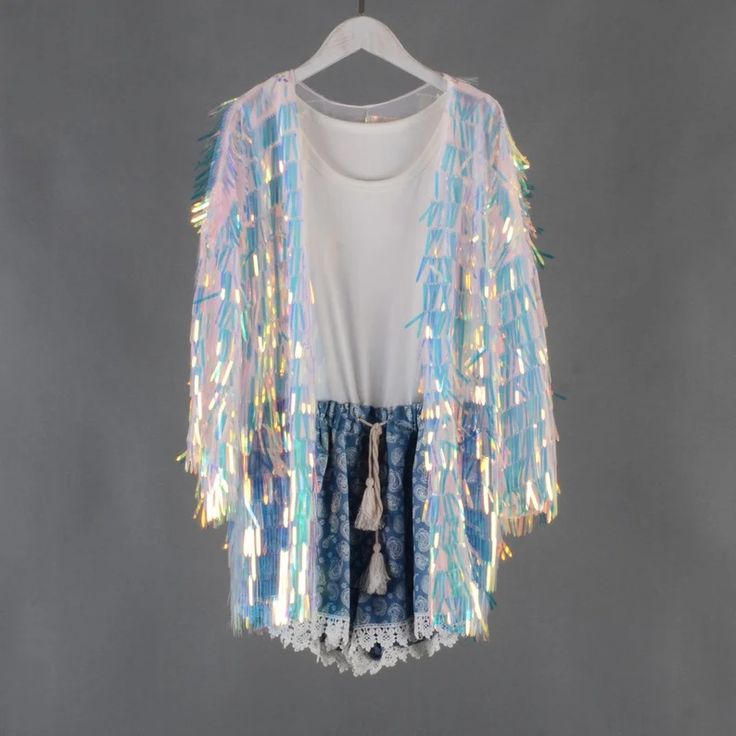 a white iridescent sequin kimono jacket with fringe and tassels. the jacket is being worn over a white tshirt and denim shorts with white lace trim. the model is standing against a light gray background.