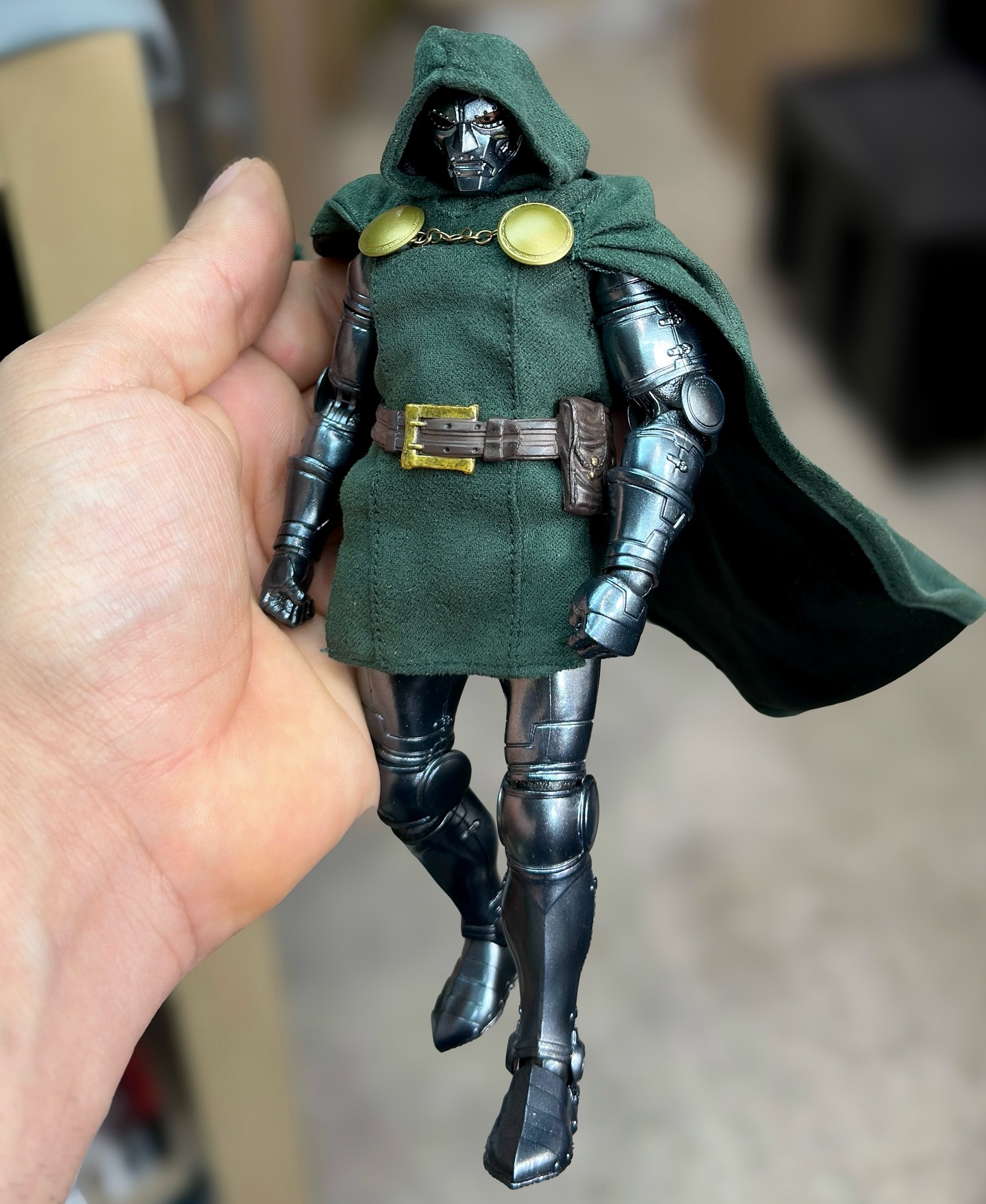 a hand holds an action figure of dr. doom. the figure is armored in metal, with a green tunic and hooded cape. the figure has a metal mask with a hood.