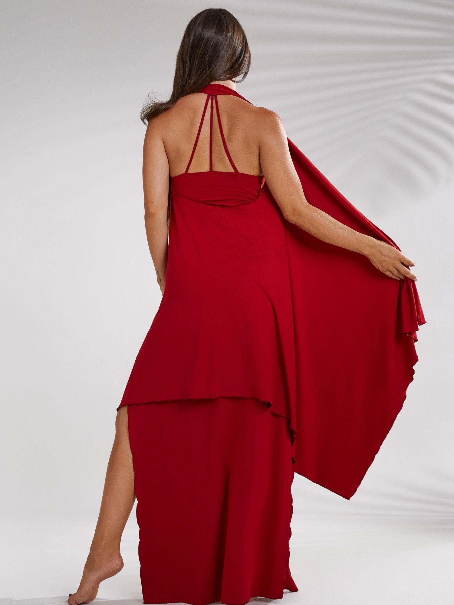 back view of a woman wearing a long, red maxi dress. the dress has a halter neckline, a high slit on one side, and a flowing, asymmetrical design. the fabric appears to be smooth and lightweight. the model is standing with her back to the camera, showcasing the drape and flow of the dress. the background is simple and uncluttered, focusing attention on the garment. the overall style is elegant and formal.