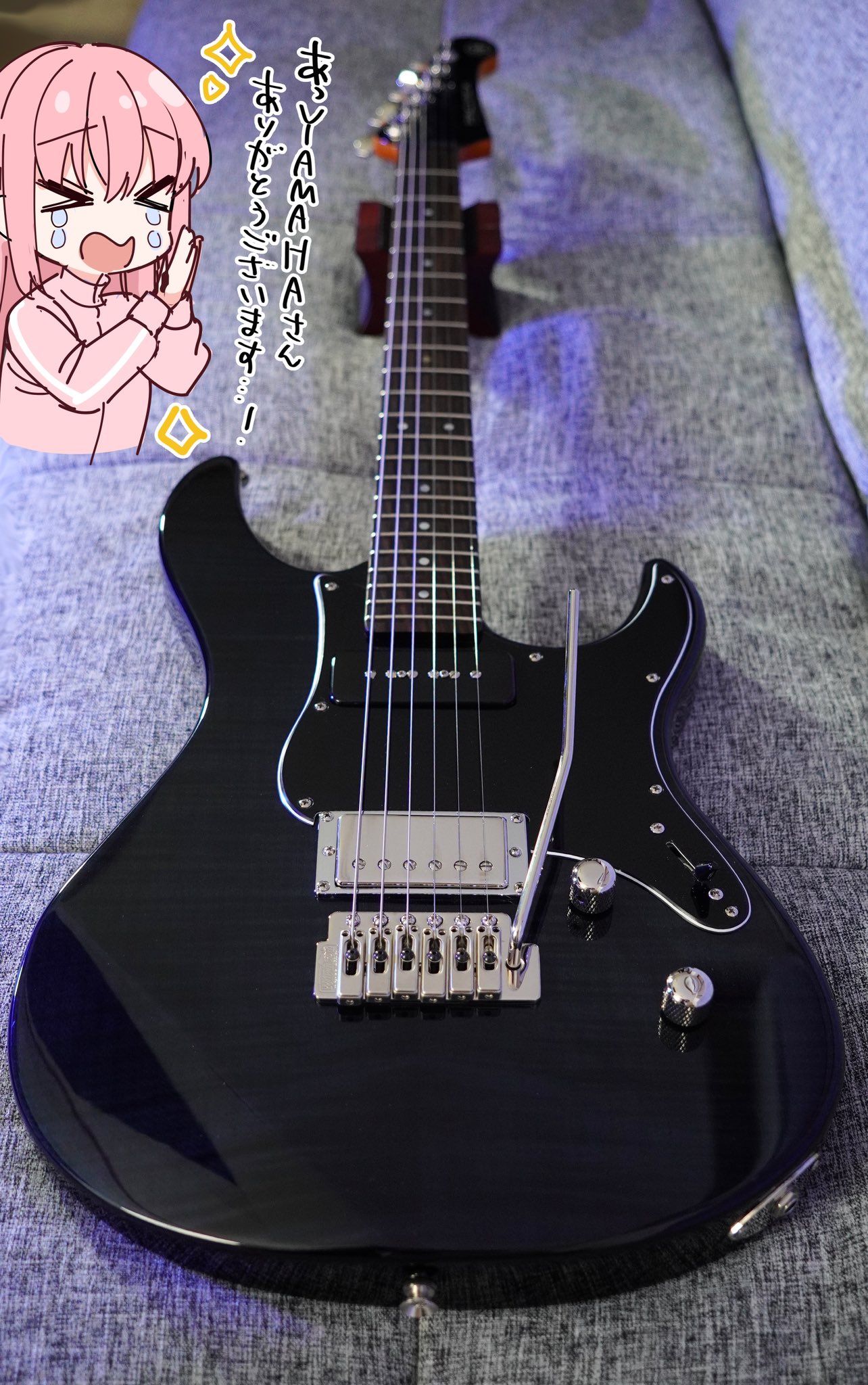 a black yamaha pacifica electric guitar is placed on a grey cushion, with an anime character expressing gratitude towards yamaha, indicated by japanese text in the image. the guitar is on a guitar stand. the focus is on the instrument and its features, such as the strings, bridge, pickups, and tremolo arm.