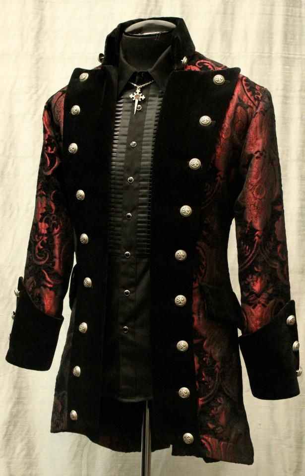 a gothicstyle black and red coat is displayed on a mannequin. the coat is dark and has a patterned red design on the sleeves and parts of the body, with black velvet panels down the front. it features numerous silver buttons and a black collar. underneath the coat, there is a black shirt with a ruffled front and a silver cross necklace. the mannequin is set against a neutral background.