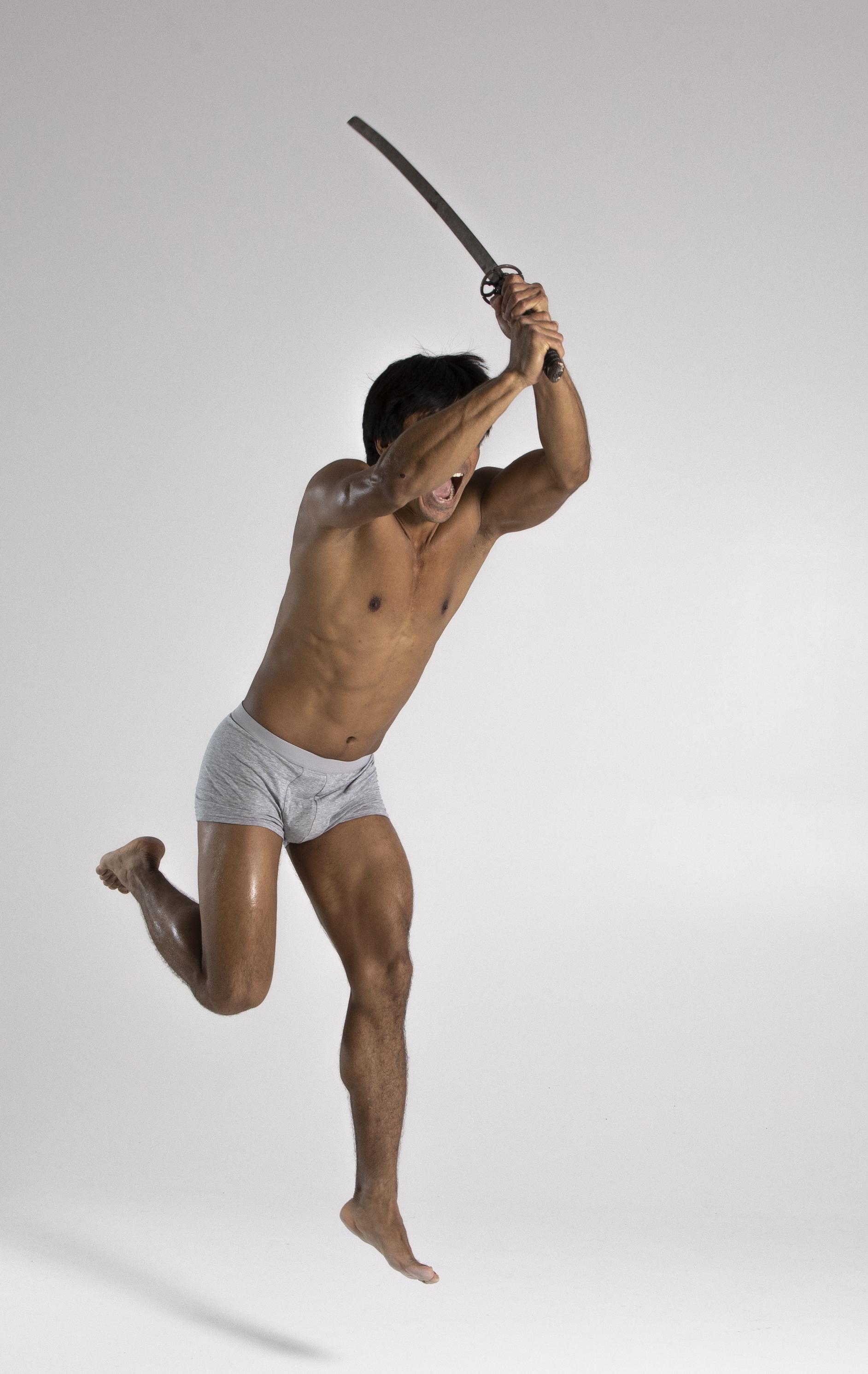 a shirtless, muscular man wearing gray underwear is jumping in the air while wielding a katana above his head in a dynamic, aggressive pose. the man has a dark complexion and is barefoot. his expression appears intense and fierce, as if he is engaged in combat. the background is a plain white or light gray.