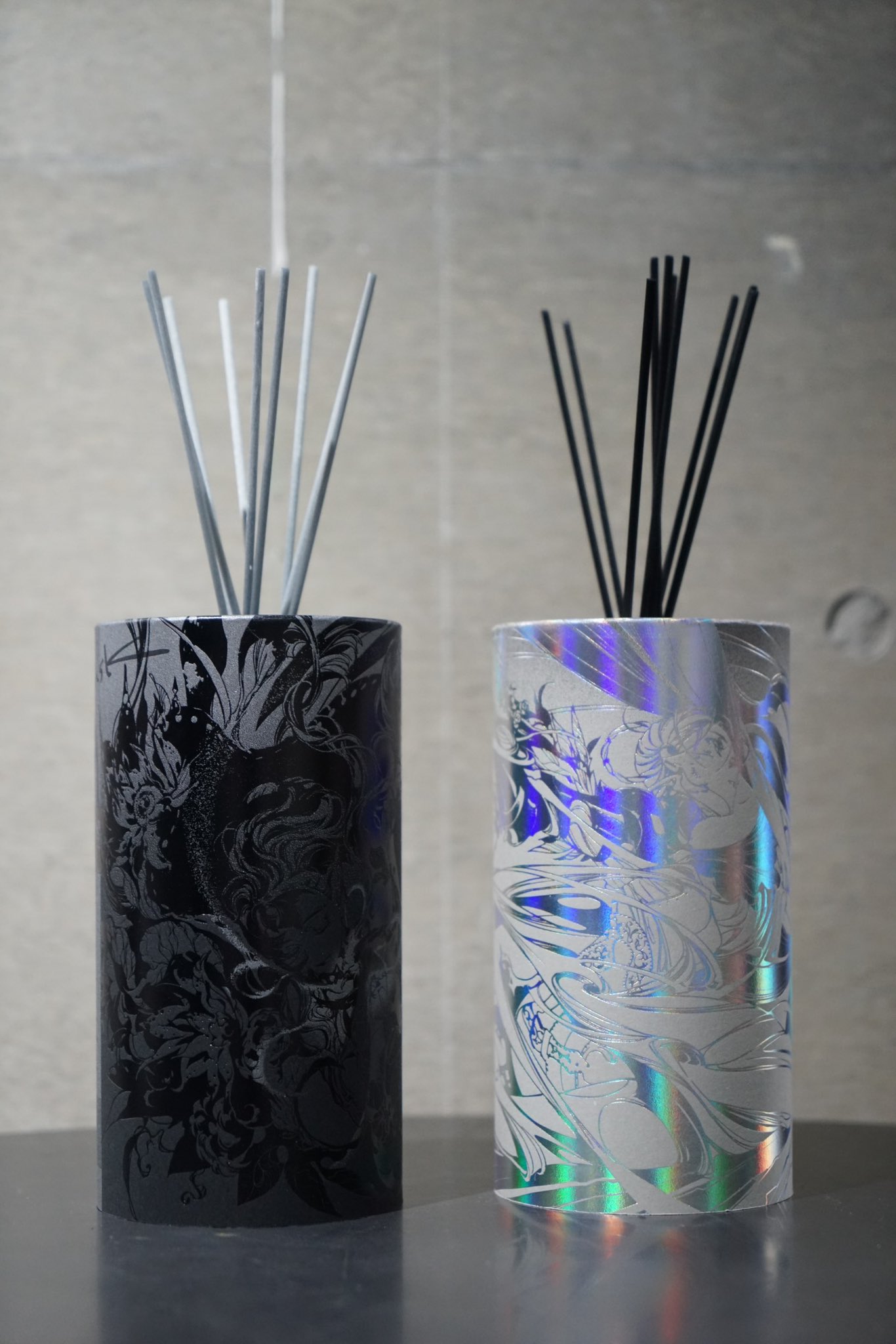 the image shows two reed diffusers on a dark gray surface in front of a gray wall. the left diffuser features a black and gray design, while the right diffuser has a white and holographic design. the background is out of focus, drawing attention to the two diffusers.