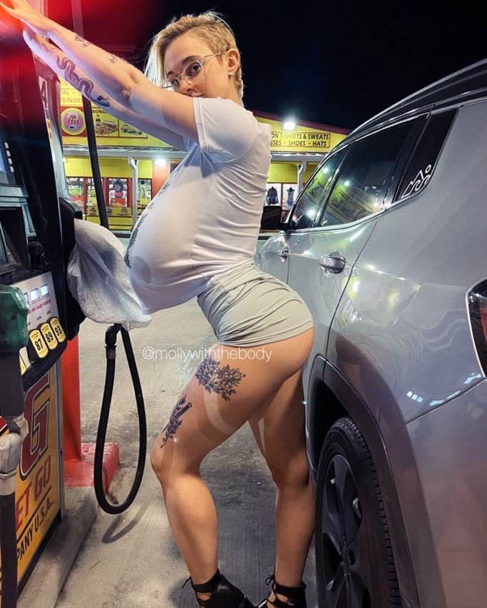 the image shows a young blonde woman with short hair and several tattoos on her legs. she's wearing a white tshirt that's stretched taut over her large breasts and short grey shorts. she is at a gas station at night, filling a grey suv.  she's leaning against the gas pump, posing confidently.  the image is suggestive and highlights her body shape, with a focus on her physique. the instagram handle "@mollywiththebody" is visible, suggesting a possible social media context.