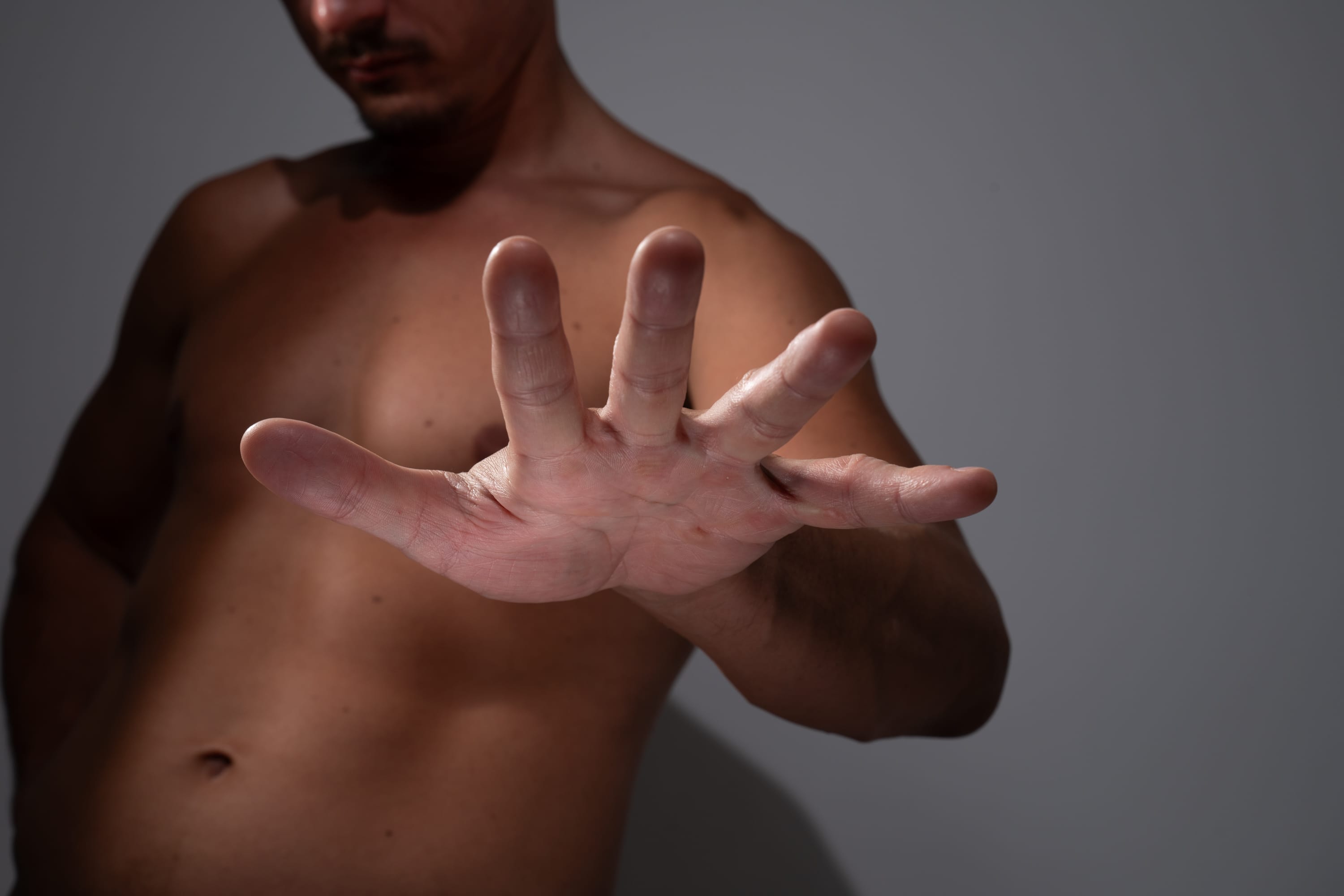 a shirtless man with a goatee stands against a gray background. his hand is extended towards the viewer in a gesture of "stop" or rejection. the image is lit with dramatic side lighting, creating strong shadows and highlights on his body.