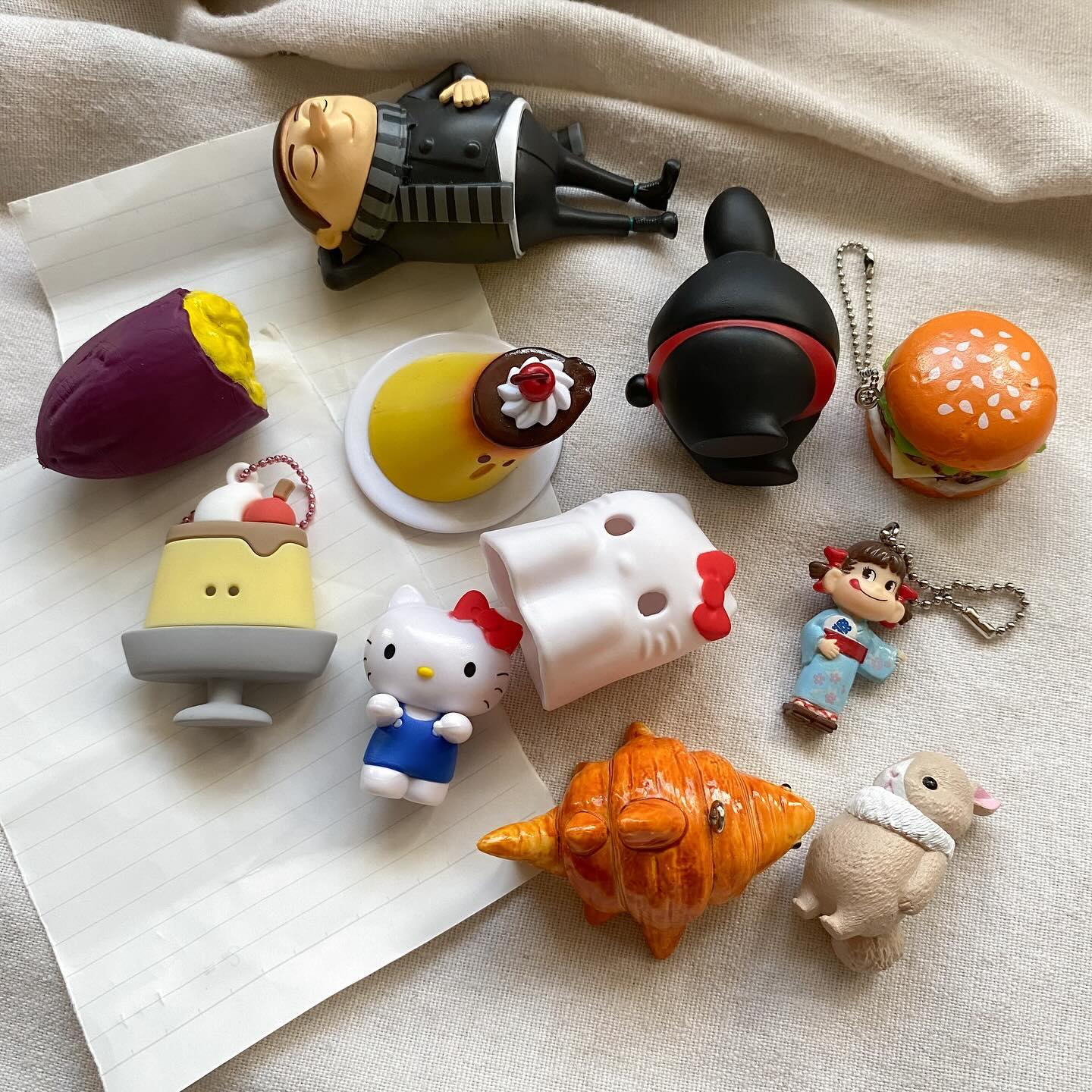 the image contains a collection of miniature figurines and toys arranged on a surface. there is a gru figure lying down, a sweet potato, two different pudding figures, a hello kitty figurine, a daruma doll, a hamburger keychain, a girl figure, a rabbit figurine, and a croissant figurine. they are arranged in a seemingly random order, creating a playful and diverse composition.