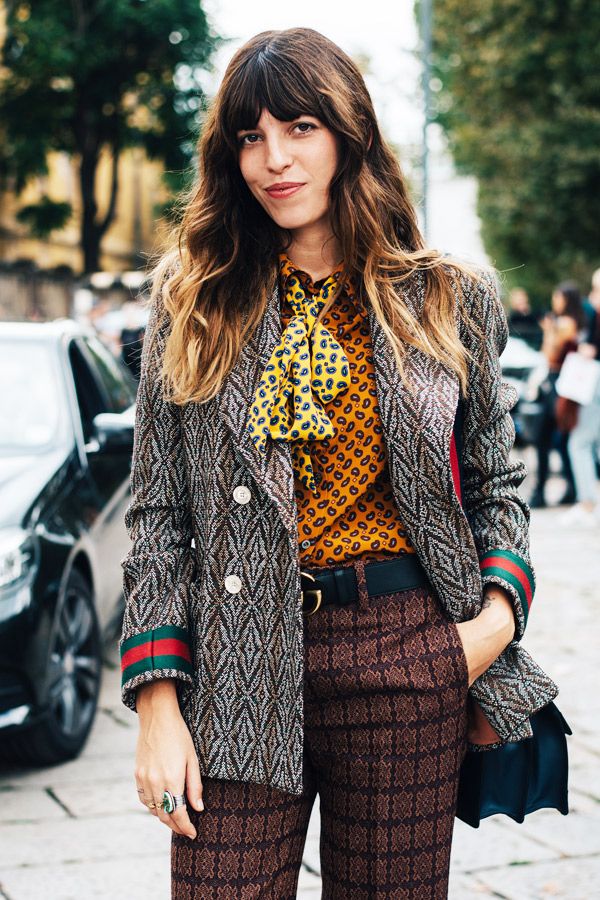 a stylish woman with long brunette hair and bangs is captured in a street style portrait. she's wearing a gray patterned blazer with green and red stripes on the cuffs, a brown and gold patterned shirt, a yellow and brown patterned scarf tied in a bow, and brown geometric patterned pants. a black belt cinches her waist.  she accessorizes with rings. a portion of a darkcolored handbag is visible.  the backdrop is a city street.