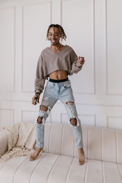 a young man with long brown hair is playfully jumping on a beige couch in a living room. he is wearing a light brown cropped sweater and light blue ripped jeans. he's barefoot and has a happy, joyful expression on his face.  the background features white paneled walls.
