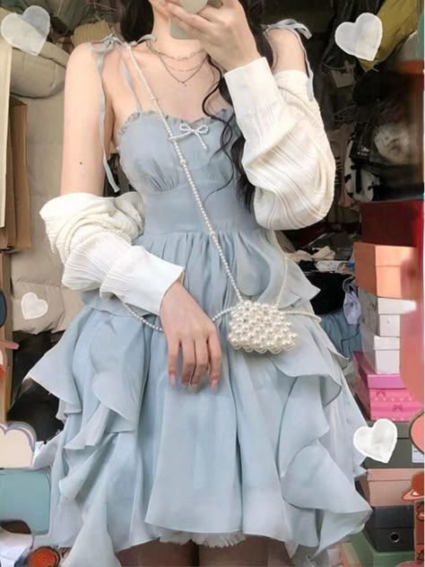 a young woman in a light blue dress with white ruffle details poses for a photo in an indoor setting. the dress features thin straps and a light blue, ruffled skirt. the woman is also wearing a white cardigan over the dress, with a pearl necklace and a small white purse. the background of the photo is blurred and features a pink box and a white box with a heart on it.