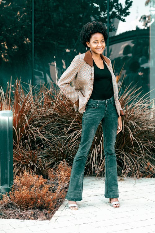 a young african american woman stands in front of a modern glass building. she's smiling and wearing a stylish outfit: a brown checked blazer over a black top, and darkwash bellbottom jeans.  she is accessorized with simple earrings and sandals. the setting is outdoors, with landscaping visible behind her. the image is a full body portrait showing her from the knees up.
