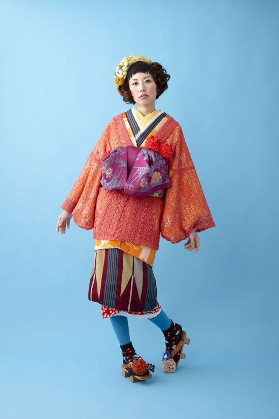 a woman is standing against a light blue background, wearing a colorful and unique kimonostyle outfit. she has short, dark hair and is wearing a floral headpiece. the kimono features an orange lacelike outer layer over a colorful patterned fabric. a purple obi with floral embroidery is tied around her waist. she is also wearing blue tights and wooden geta sandals with colorful socks. her expression is neutral.