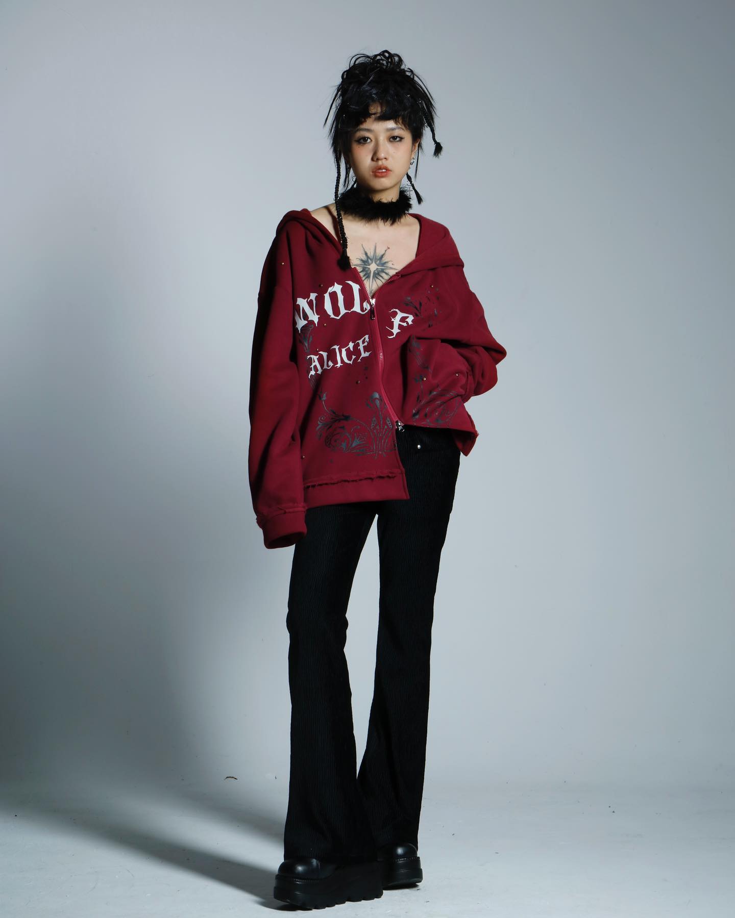 a young woman with dark hair styled in an updo poses in a studio against a light gray background. she's wearing a dark red oversized zipup hoodie with white graphic text that partially reads "alice," black bellbottom pants, and chunky black platform boots.  she also wears a black choker necklace.  her overall style is edgy and alternative.