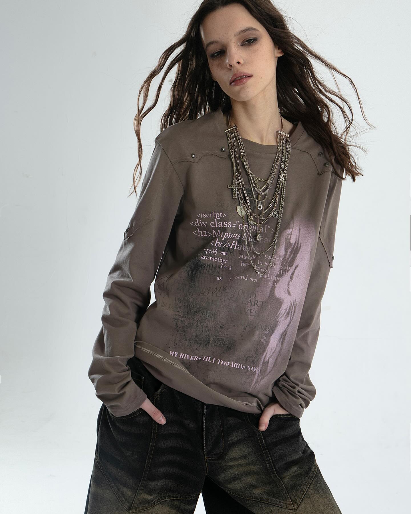 he image shows a female model wearing a taupe longsleeved shirt with a graphic print that includes both text (seemingly code and poetic text) and an artistic image.  the shirt has a distressed look, particularly around the shoulders. she is also wearing several layered silver necklaces featuring crosses and other small charms. the model's bottom half is visible, showcasing darkwash, baggy, distressed denim pants. her long brown hair is slightly tousled. the overall style is grunge, casual, and incorporates elements of alternative and streetwear aesthetics. the background is a simple, neutral light gray.