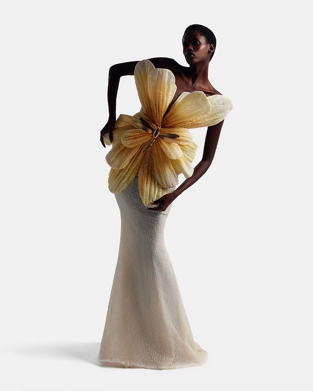 a black woman wearing a stunning couture dress. the dress is white and has a large yellow flower on the top half. she is standing in a studio with a simple white background.