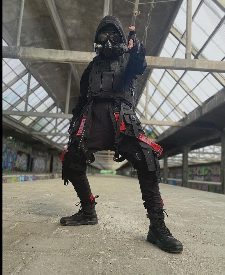 a man in a black outfit and gas mask stands on a gritty concrete surface, posing for a photo shoot. he's wearing black pants, boots, and a jacket. he has many red straps on his legs. he's looking intently at the camera,  as if he's a tough character in a postapocalyptic film.