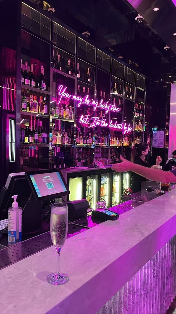 a vibrant bar scene bathed in pink neon light. a neon sign reads "you may be my lady, but i'm the alchemist, baby". the bar is stocked with liquor bottles and a champagne flute sits on the counter. a bartender is partially visible in the background.  the ambiance suggests a lively and stylish nightlife setting.