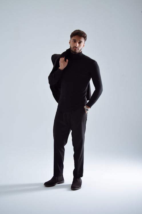a man is standing in a studio wearing a black turtleneck, black suit, and black shoes. he is posing with a suit jacket over his shoulder. the background is light gray.