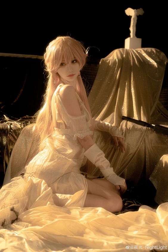 a woman is posing in a white dress with a golden wig. she has horns on her head, suggesting she is cosplaying as an anime or manga character. the setting appears to be a bedroom or studio with golden drapes and warm lighting.