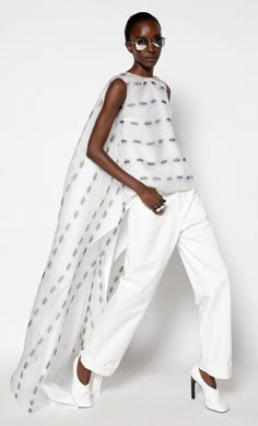 a fashion model wearing a white dress with a white patterned overlay and white pants. she is also wearing white shoes, sunglasses and has her hair pulled back. she is posing with her hands on her hips, and she is standing in a studio on a white background. the image is a portrait shot from the waist up.