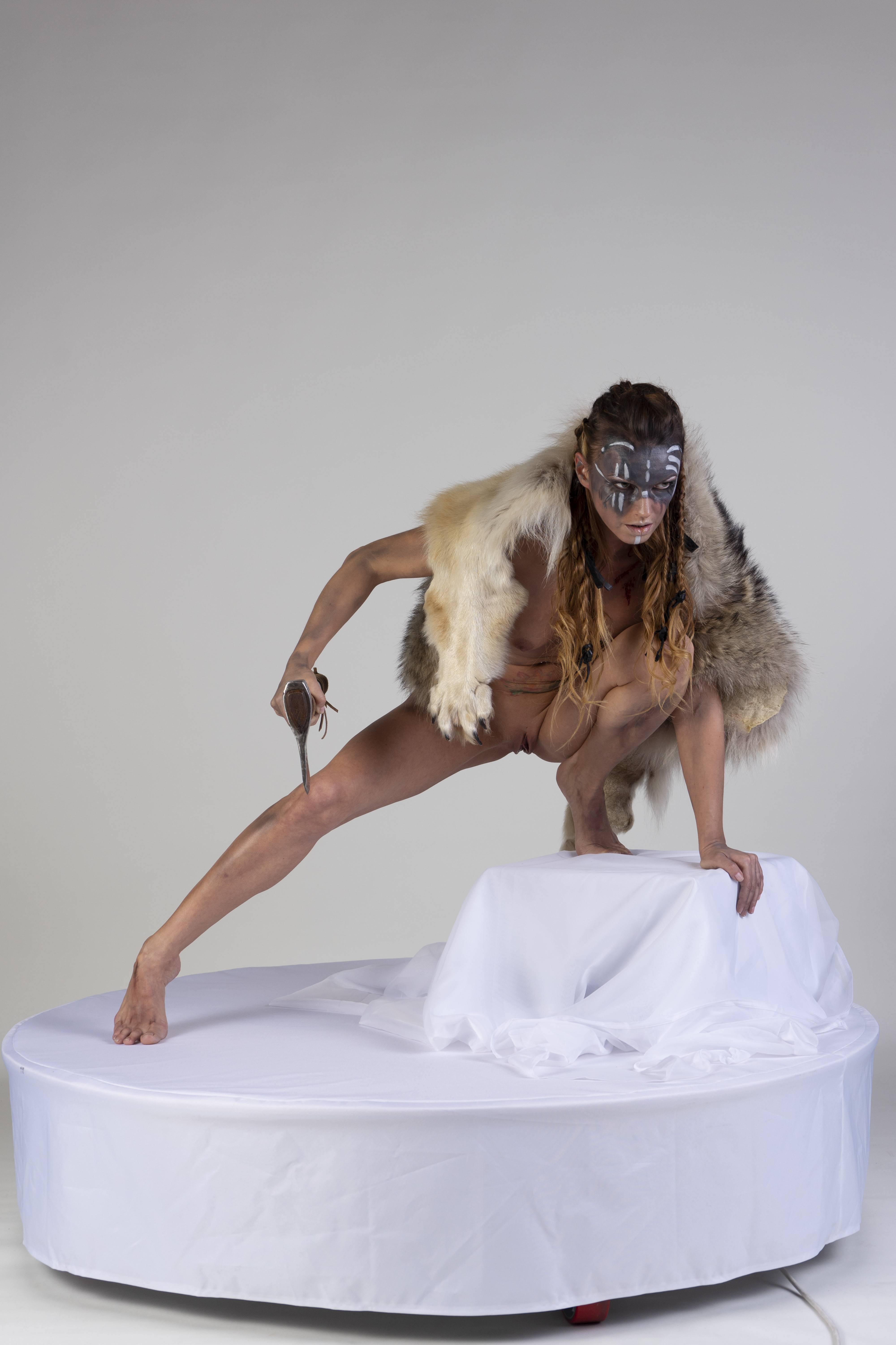 a woman with a fur vest and a painted mask is crouching on a white platform, holding a spear. she is positioned against a white background in a studio setting.