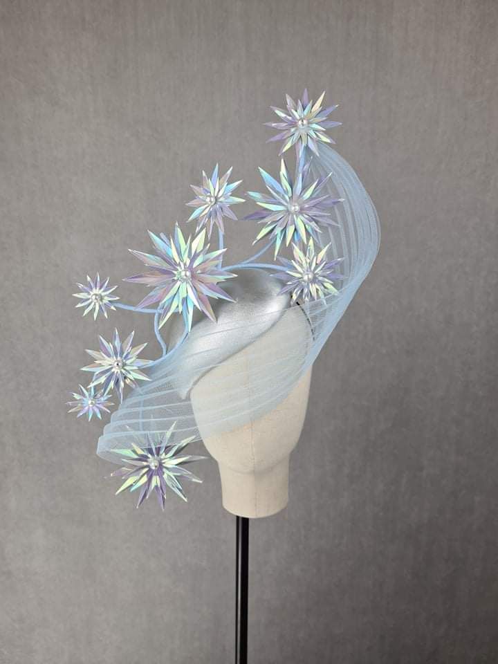 a fascinator headpiece sits on a mannequin head, styled against a gray backdrop. the headpiece is a mix of silver and light blue tulle fabric, adorned with iridescent starburst accents. the stars vary in size and are positioned to create a dynamic and visually striking effect.