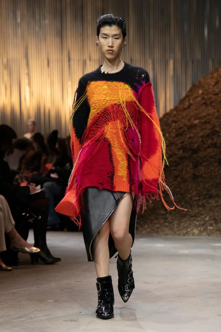 a woman in a colorful sweater and leather skirt walks down a runway. the sweater is red, orange, and yellow with black accents and has long, frayed ends. the skirt is black leather and is wrapped around her hips. she is wearing black boots with silver buckles. the background is a blurry shot of people and a pile of dirt.