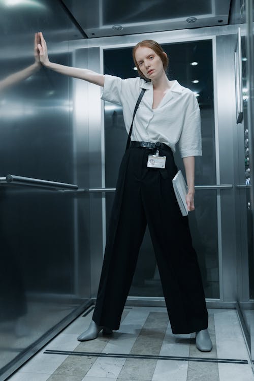 a stylish woman in a white shirt and black pants leans against the wall of a modern, metal elevator. she is holding a briefcase and has an id card hanging from her belt. she's looking towards the camera. the overall aesthetic is clean and minimalist.