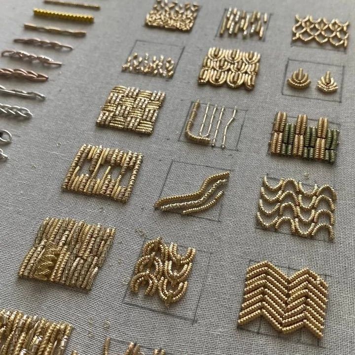 the image shows examples of goldwork embroidery techniques. various geometric patterns are created on a grey fabric base using gold and other colored metal threads. the patterns include lines, curves, zigzag shapes, and woven effects within square outlines. a selection of goldwork materials like bullion wire, beads, and couching threads are displayed on the side.