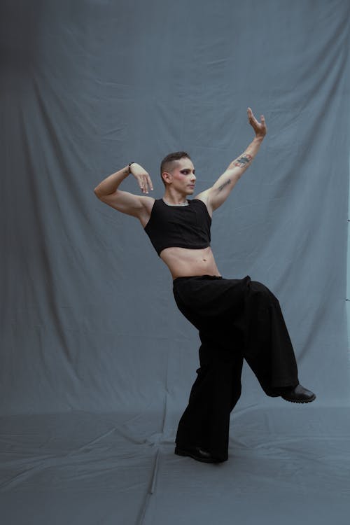 a fullbody shot of a male dancer in a contemporary dance pose. he is wearing a black crop top and loosefitting black pants.  his body is arched and his arms are extended in a fluid motion. he is barefoot, in a dance studio with a gray backdrop. the overall image is artistic and evocative of movement and grace.