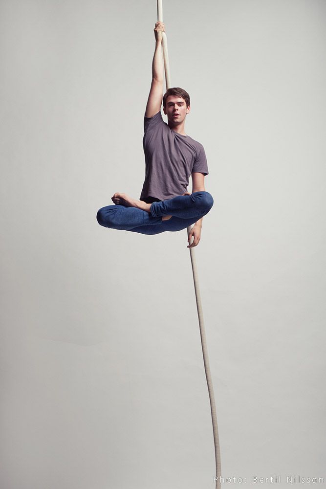 a man hangs from a rope in a gray studio. he is wearing a gray tshirt and blue jeans. he is holding the rope with one hand and his legs are crossed at the knee. the man is looking down at the camera.