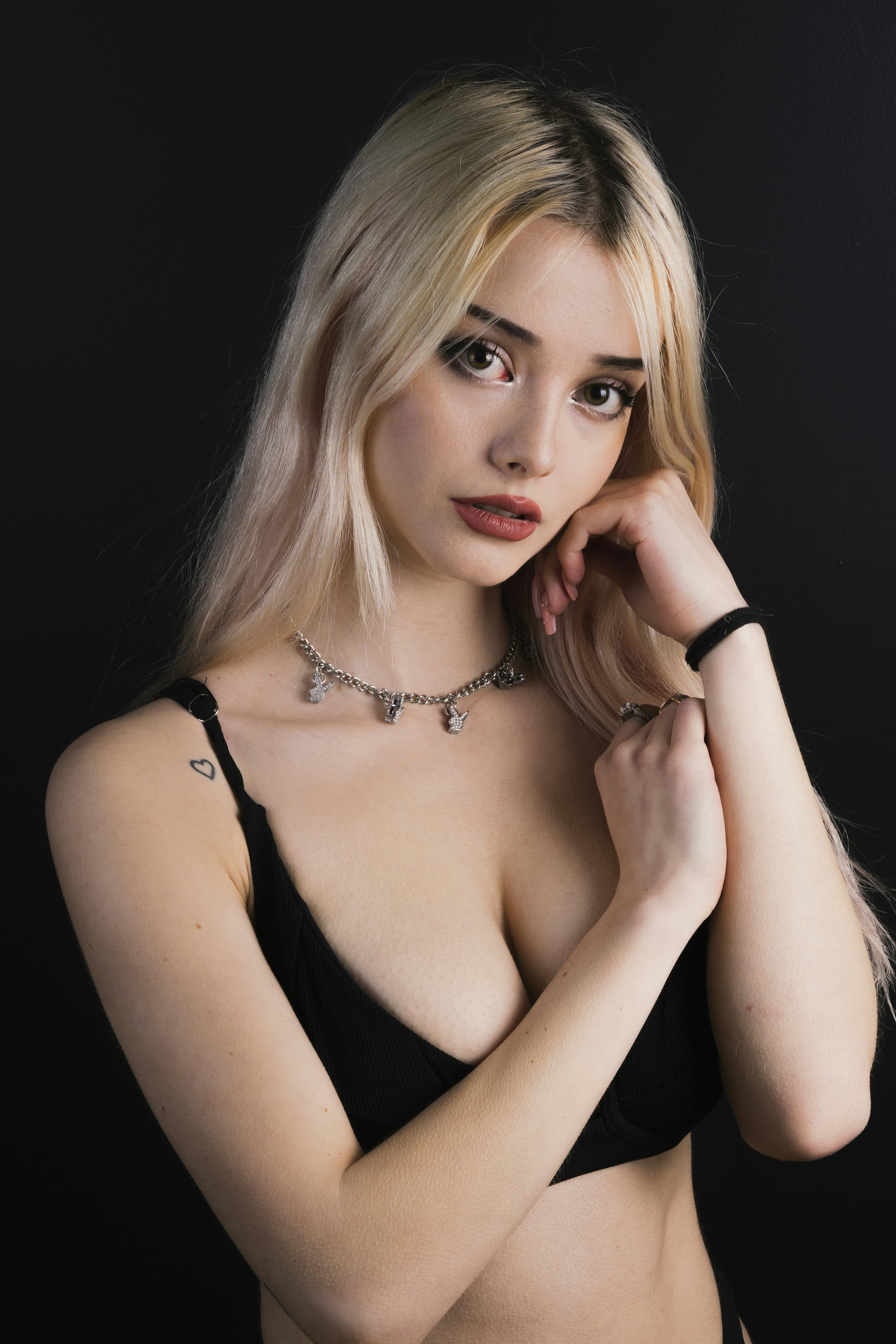 closeup portrait of a young, attractive blonde woman with long hair. she is wearing a black bra and a silver necklace with small charms. her arms are crossed over her chest, and she is looking directly at the camera. she has a small heart tattoo on her left shoulder. the background is black.