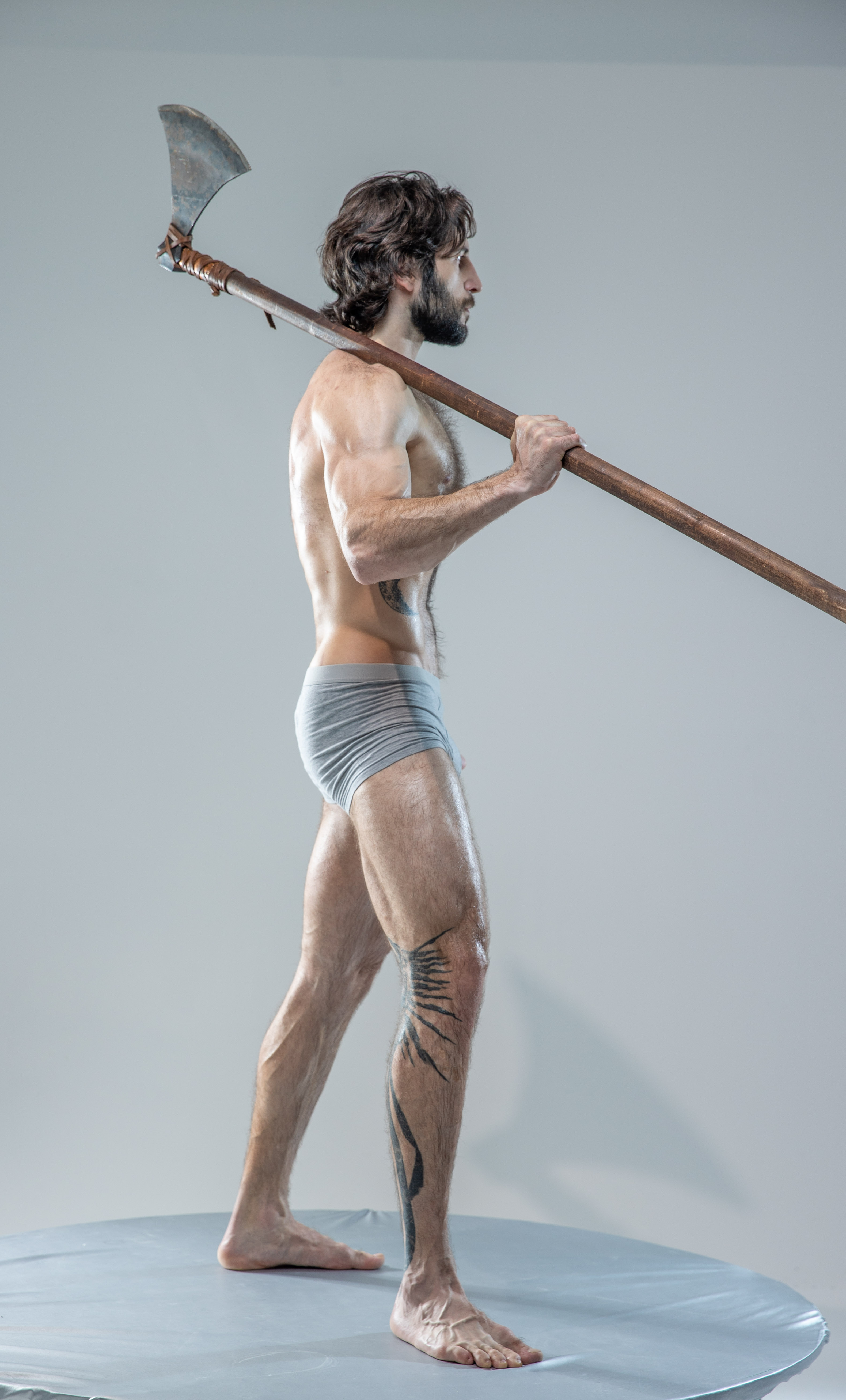 a muscular man with long hair and beard, clad only in grey underwear, stands on a round platform in a studio setting. he holds a large, doubleheaded axe across his shoulders, his grip firm and gaze intense. his pose is strong and evokes a warriorlike presence. the lighting is soft and highlights the definition of his muscles.