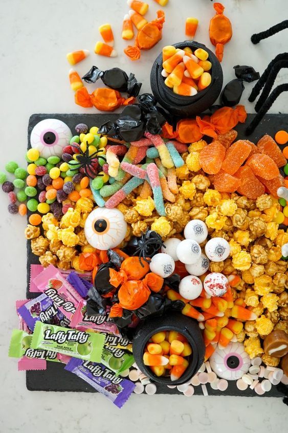 a detailed closeup shot of a halloween candy board overflowing with a wide variety of sweets.  the board is rectangular and dark gray, acting as a backdrop for the vibrant candies arranged in seemingly random yet visually appealing clusters.  different types of candies are identifiable, including gummy worms, candy corn in small black cauldrons, popcorn, various jelly beans, eyeshaped candies, orange slices, laffy taffy candy bars, and chocolate candies.  small plastic spiders and other halloween decorations are interspersed amongst the candy. the board rests on a light marblelike surface.  the overall impression is one of abundant, festive halloween treats ready for a party or celebration.