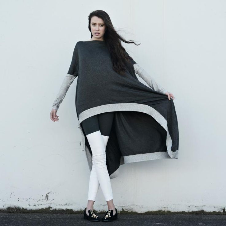 a full shot of a woman posing in front of a white wall, wearing an asymmetrical dark gray top with light gray trim, longsleeved light gray underlayer, black and white leggings, and black shoes. she is holding the edge of the top to showcase the design. her long dark hair is flowing in the wind.