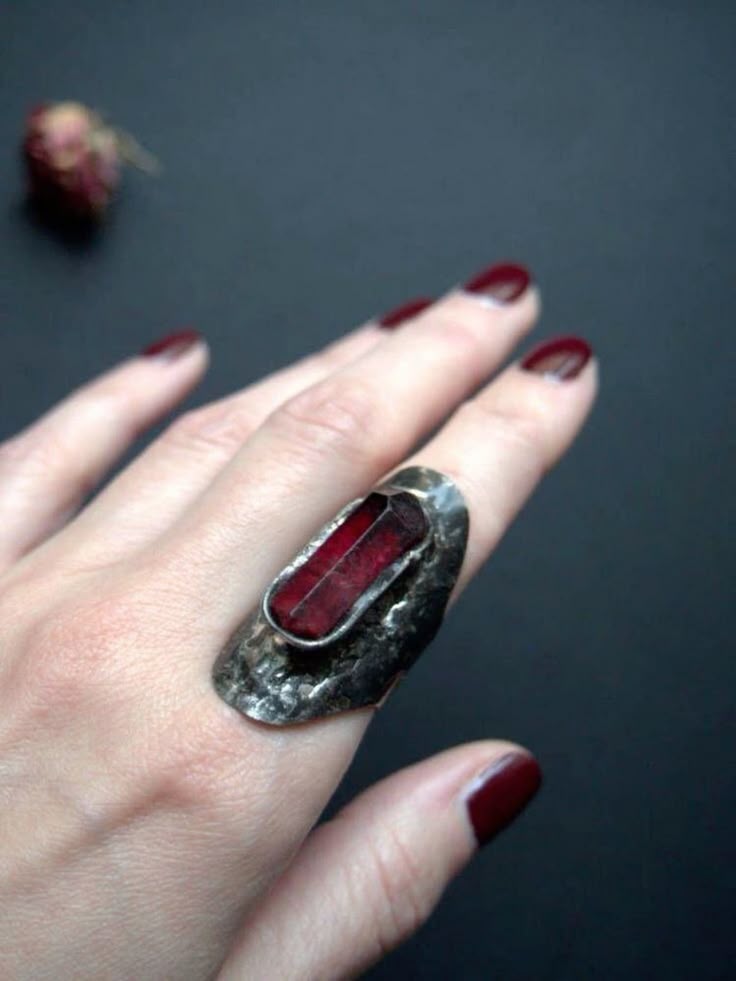 a hand with dark red nail polish on the fingers is wearing a large silver statement ring with a red stone on the ring finger. a dried rose bud sits in the background. the background is black.