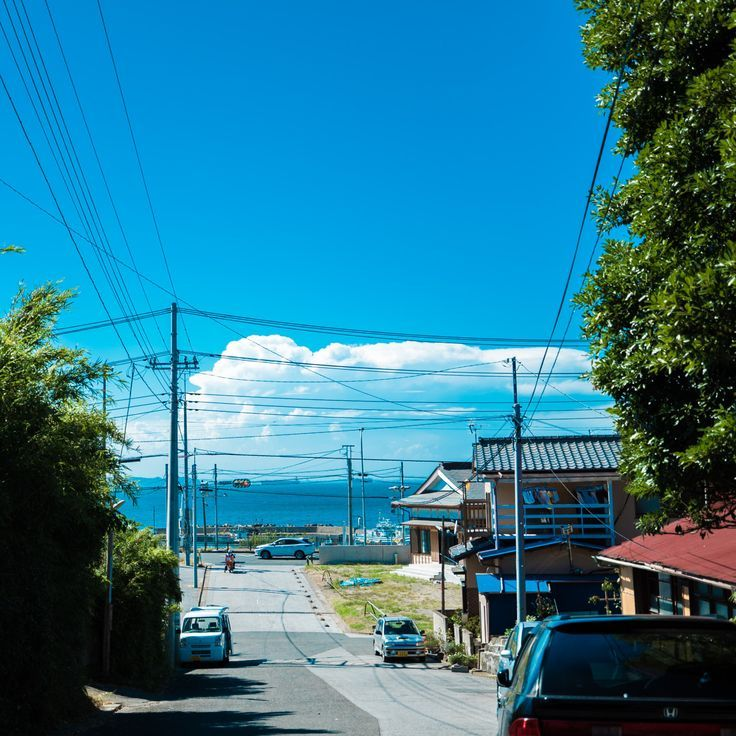 this image captures a street view in a residential neighborhood on a sunny day. the road slopes slightly downhill towards the horizon, where the sky meets the sea. multiple cars are parked or moving along the street, with houses and trees lining both sides. the sky is a clear blue with some clouds, and utility poles with electrical wires crisscross above the street.