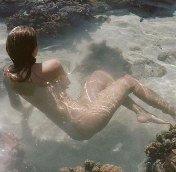 a woman is lying on her back in shallow water surrounded by coral reefs. the sunlight streams through the water, creating an ethereal effect on her body.