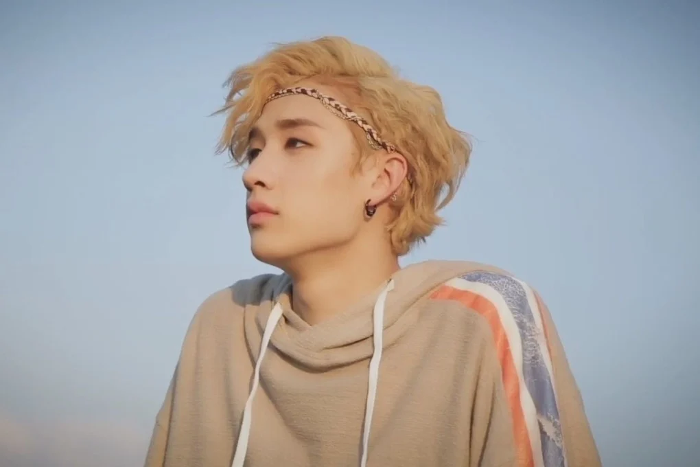a portrait of a young man with blond hair, looking to the right, with a relaxed and casual expression. he is wearing a tan hooded sweatshirt, a headband, and an earring. the backdrop is a blue sky.