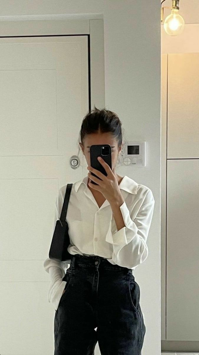 a woman is taking a selfie in a bathroom mirror. she is wearing a white buttonup shirt and black pants. she has a black shoulder bag hanging off her left shoulder. her hair is pulled back. the background includes a door, a light fixture, and other bathroom fixtures. the overall style is minimalist and clean.