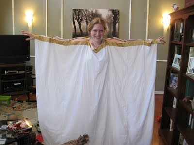 a woman stands indoors, holding a large white sheet or robe with golden trim. she is smiling. behind her is a living room setting with a television, bookshelf, and framed picture on the wall. the floor appears to be messy.