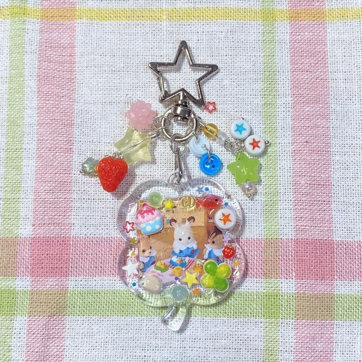 a closeup of a decorative keychain on a plaid background. the keychain features a starshaped keyring at the top, from which hangs various charms and beads, including a strawberry, pink flower, and small colorful buttons. below this, there's a clovershaped clear plastic charm filled with tiny animal figures, a miniature cupcake, and other small objects. the keychain is set against a background of a pink, green, and white plaid cloth.