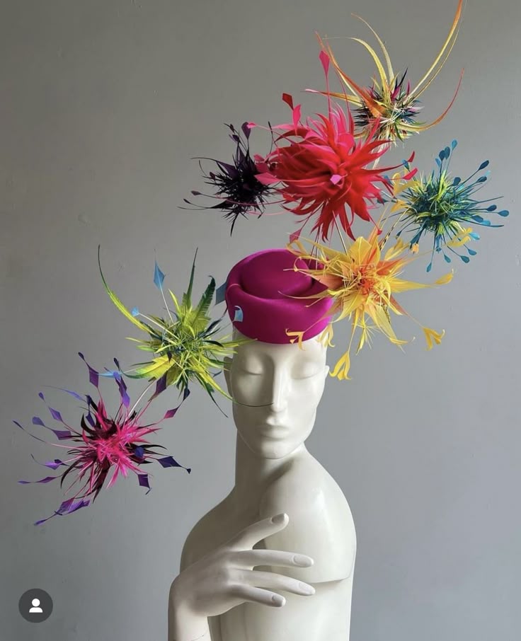 a brightly colored, elaborate hat or headpiece is displayed on a white mannequin. the hat is magenta with various colorful, feathery decorations extending outwards, creating a spiky, floral appearance. the background is a plain gray. the mannequin's hand is placed gracefully on its chest.