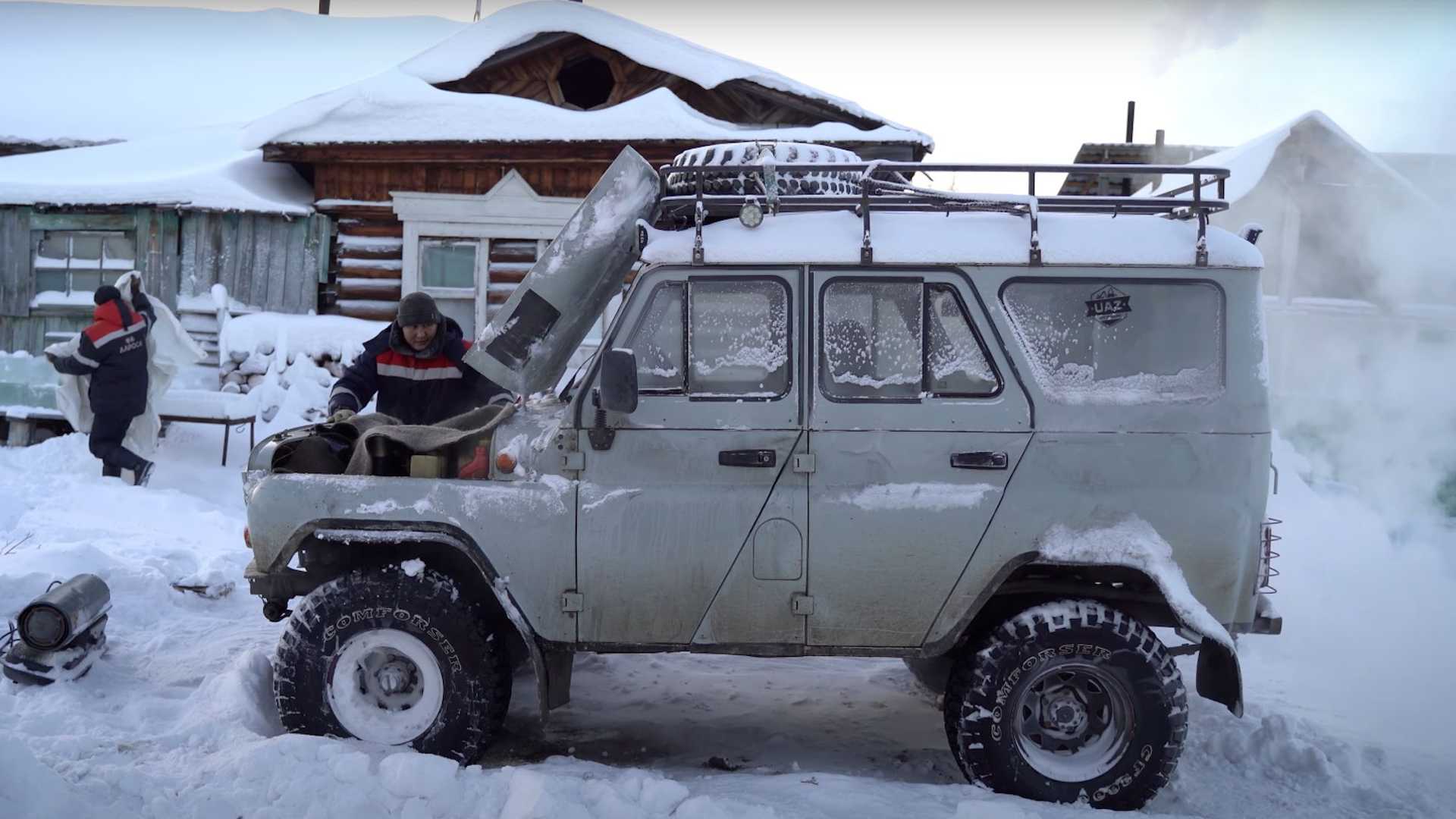 a uaz russian car, parked in a snowy, rural village during winter, with a man working on the engine of the vehicle. the car is covered in snow, and the ground is also covered in snow. there are houses in the background, and trees are visible. it is a bright, sunny day.
