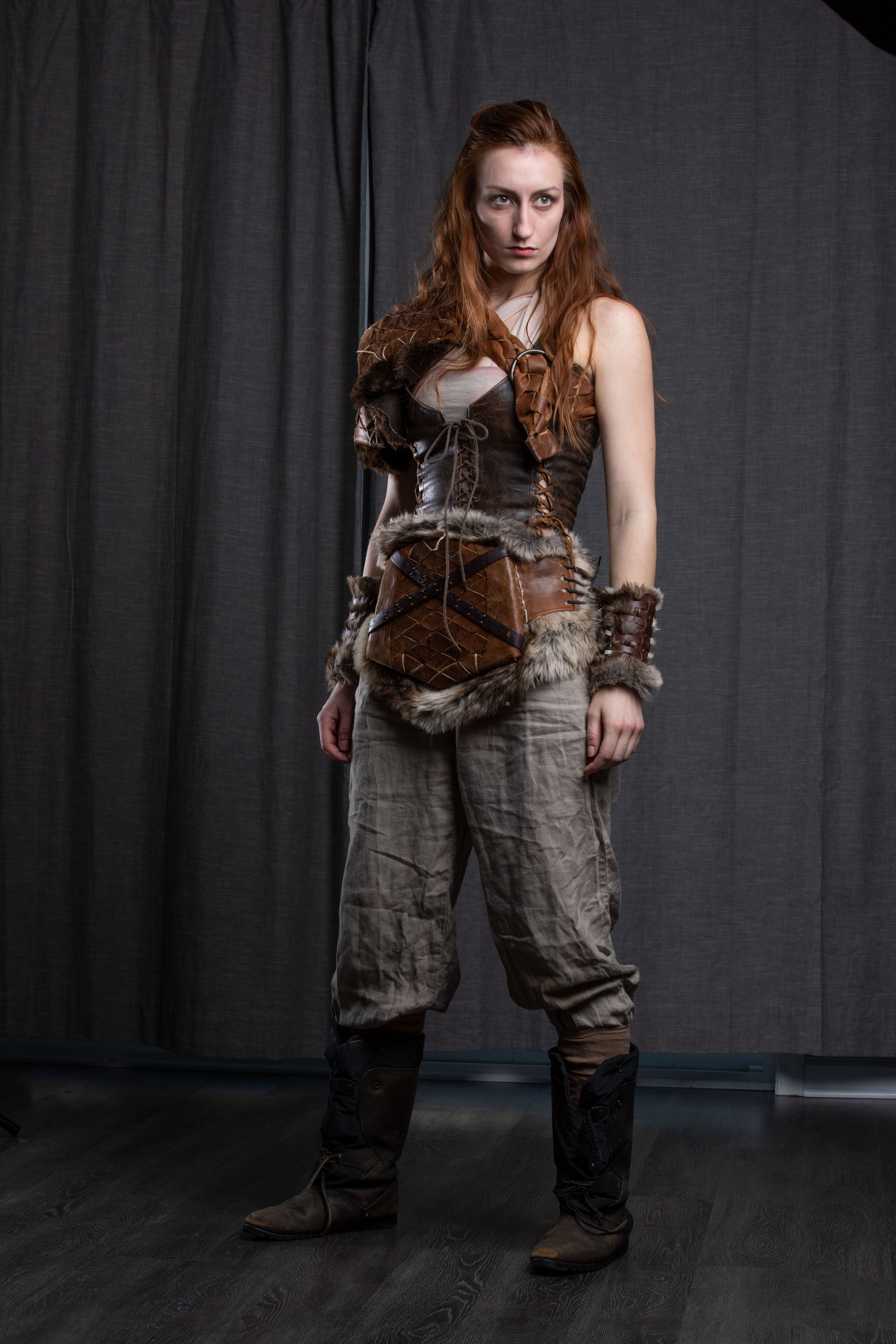 a young woman with long red hair stands against a dark grey backdrop, embodying a fierce warrior.  she wears an elaborate costume comprised of a leather bodice, furtrimmed shoulder armor, and matching wrist cuffs. her attire is completed with loosefitting linen pants and worn leather boots. the overall aesthetic evokes a sense of strength, independence, and a touch of fantasy, possibly inspired by historical or fictional warrior cultures. the lighting is soft and diffused, casting a gentle glow on the subject and highlighting the textures of her costume.