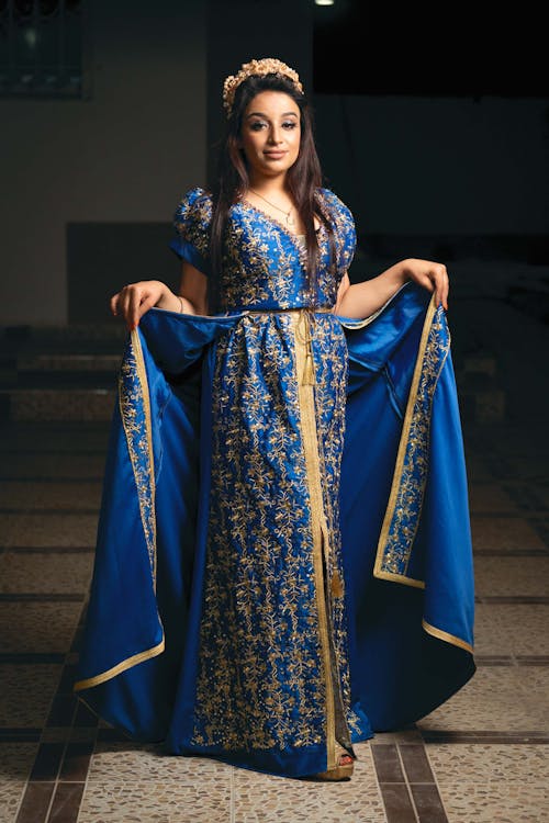 the image shows a young woman elegantly posed in a luxurious, floorlength moroccan caftan (or takchita). the caftan is a vibrant royal blue, richly adorned with intricate gold embroidery throughout.  a gold belt cinches her waist, and a matching gold headpiece adds to the regal appearance. the fabric appears to be satin or silk, creating a graceful flow.  she is positioned indoors, on a tiled floor, suggesting a formal setting such as a celebration or event. the overall impression is one of beauty, elegance, and cultural significance.