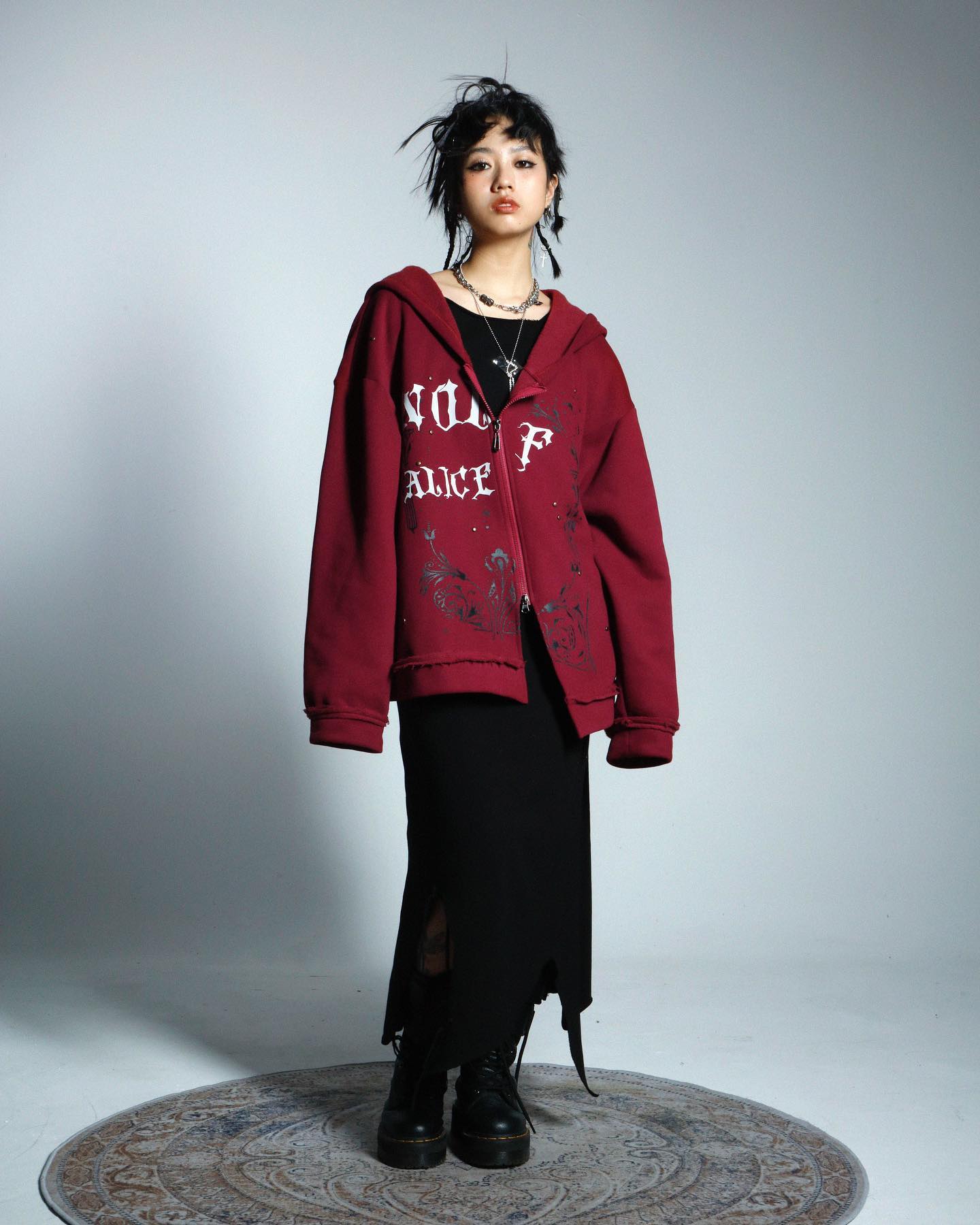 a young asian woman is posing in a studio setting against a light gray backdrop. she's wearing an oversized, dark red hoodie with "wolf alice" printed on it in a gothicstyle font, along with other subtle graphic details.  the hoodie is paired with a black, long slit skirt, and black doc martensstyle boots. she has dark hair styled slightly messy, and is wearing delicate necklaces.  the overall style is a blend of grunge, dark academia, and streetwear aesthetics. the image is welllit and emphasizes the clothing details. she is standing on a circular rug.