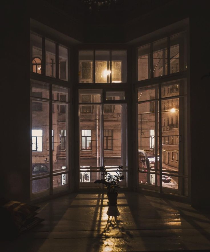 the image shows a cozy interior scene at night.  the focal point is a large bay window overlooking a city street.  the street is lit by streetlights, casting a warm glow on the buildings visible through the window.  inside, the room is dimly lit, highlighting the shadows cast by the window frame and a small potted plant sitting on the window seat. the overall atmosphere is calm, quiet, and somewhat moody. the style evokes a sense of vintage or oldworld charm. the window panes are numerous and detailed showing the city view in a grand scale. a bus or tram is visible on the street below.